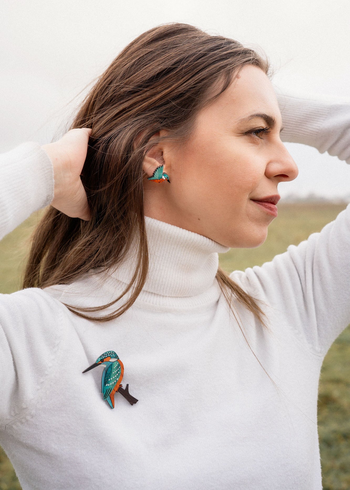 A woman in a white turtleneck stands outdoors with her hands in her hair, wearing matching colorful kingfisher bird earrings and a handcrafted Kingfisher Brooch. The background is a grassy field under a cloudy sky.
