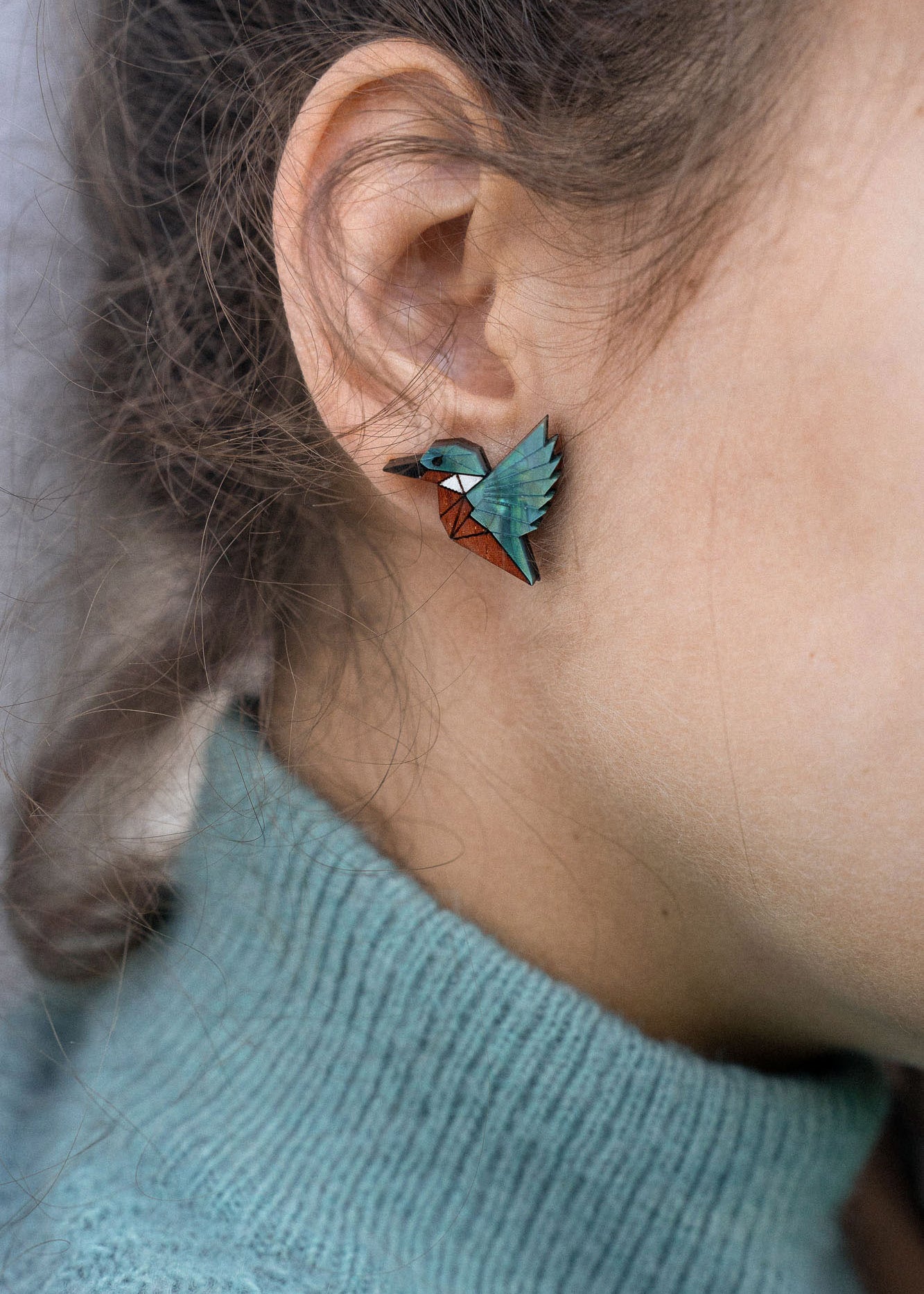 Close-up of a person wearing a turquoise sweater and eco-friendly Kingfisher Stud Earrings, shaped like a hummingbird, with the focus on their ear and earring. The background is softly blurred.