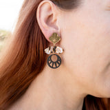 Close-up of a person with long auburn hair wearing handcrafted Kintsugi Earrings featuring a gold leaf, a clear crystal, and a black circular charm with a cut-out sunburst design inspired by Japanese art.