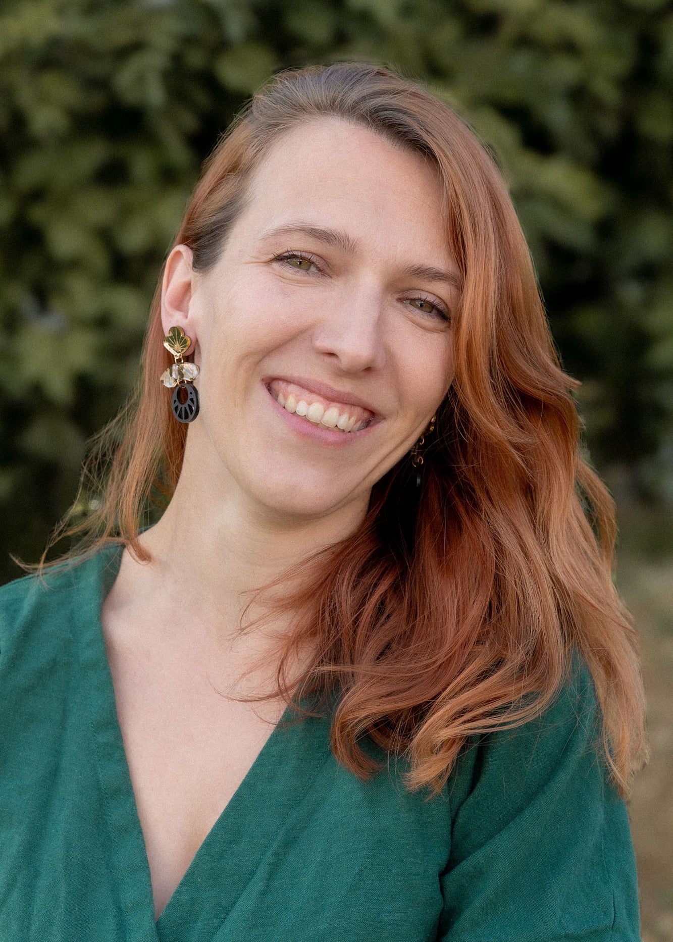 A woman with long, reddish-brown hair smiles warmly at the camera. She is wearing a green top and striking handcrafted Kintsugi Earrings. The blurred greenery in the background suggests an outdoor setting.
