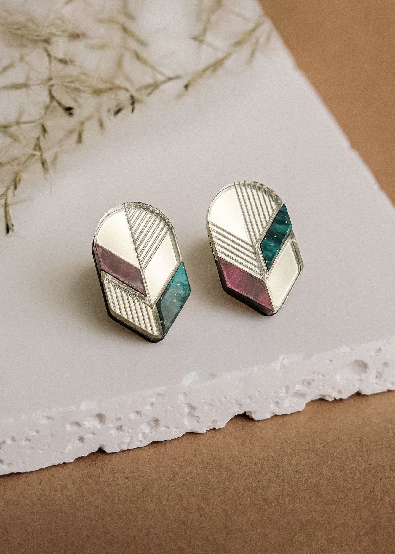 A pair of geometric stud earrings featuring silver edges and sections of white, mauve, and teal—beautifully displayed on a white textured slab with a brown background and dried plant decor—perfect for lovers of minimalist jewelry.