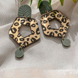A pair of Leopard Earrings featuring timeless patterns—leopard print hexagons with green leaf-like and circular accents—crafted from premium materials, displayed on light fabric with a fern leaf above.