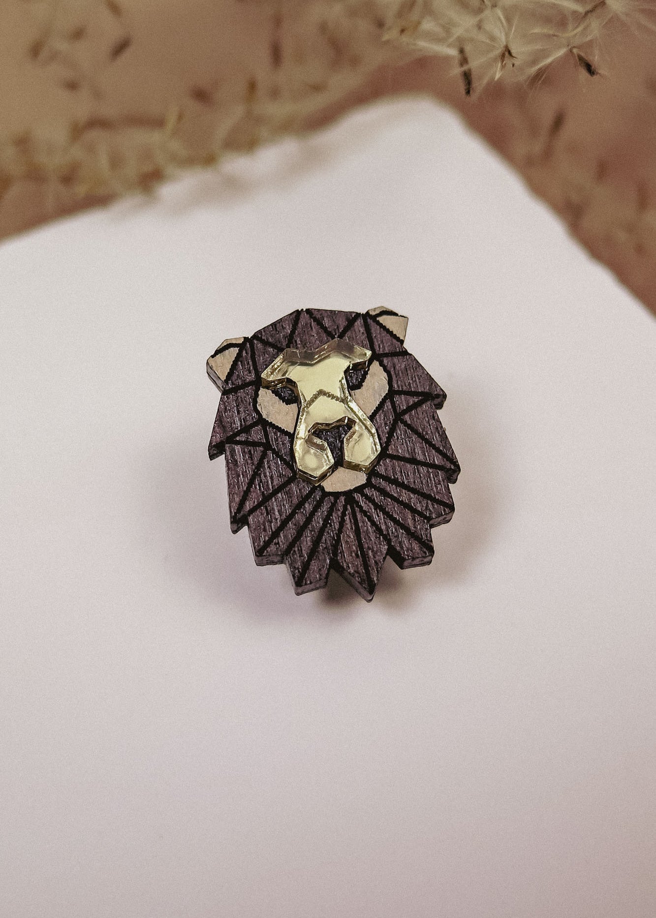 A handcrafted geometric Lion Pin made of dark wood with gold highlights, placed on a white surface with dried grasses in the background.