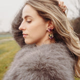 A woman with wavy blonde hair, wearing a fluffy gray coat and playful Mermaid Tail Earrings, stands outdoors with her eyes closed and one hand in her hair. The background is grassy with blurred trees—perfect for mermaid lovers.