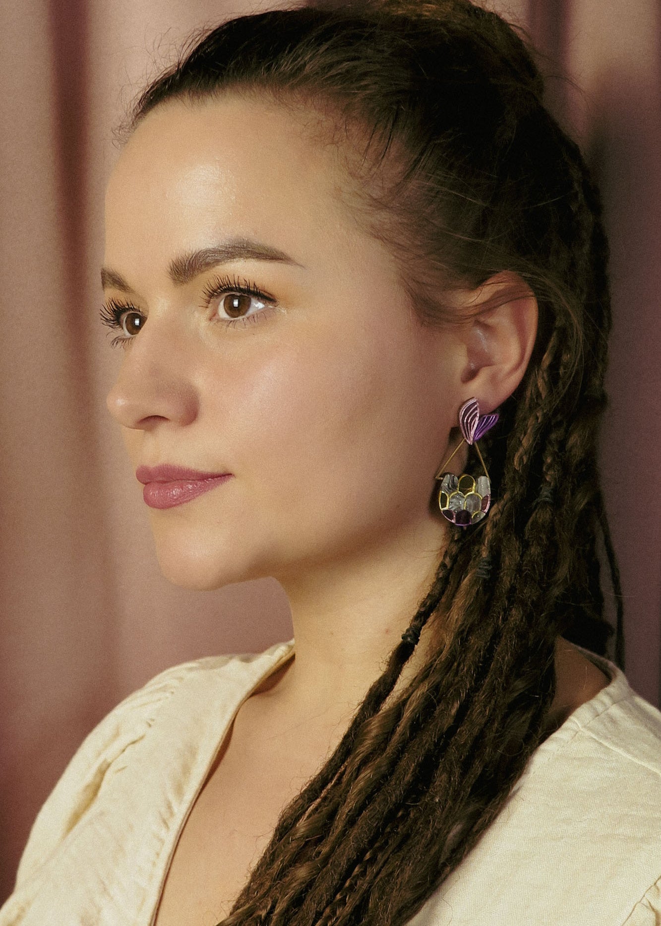 A woman with long brown dreadlocks and natural makeup looks to the side, embodying whimsical elegance. She wears a cream-colored top and geometric Mermaid Tail Earrings with purple accents. The background is softly blurred with pinkish curtains.