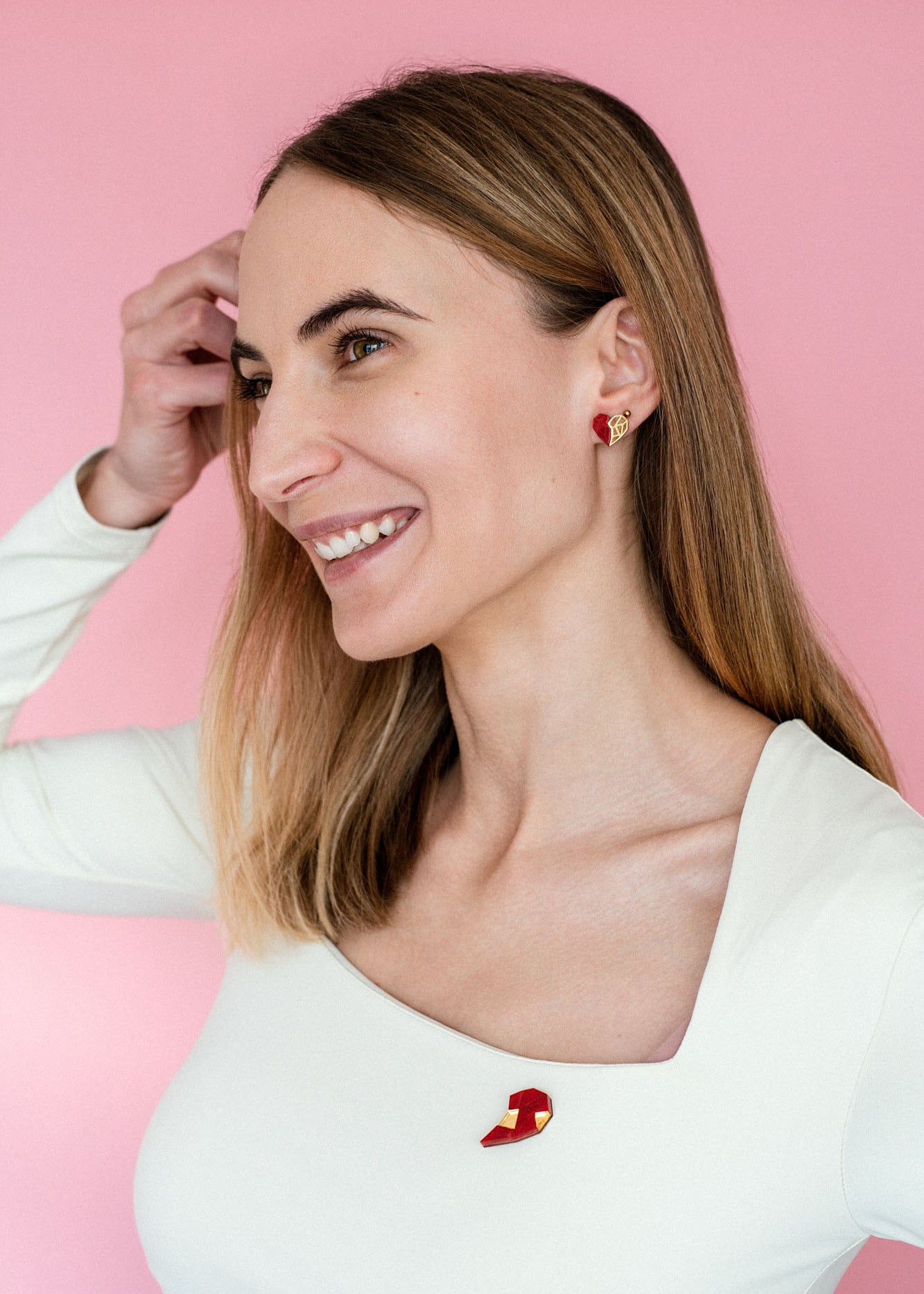 A smiling woman with straight, light brown hair wears a white, square-neck top and romantic jewelry—handcrafted earrings and a matching pin in red-and-gold geometric shapes—against a pink background. She raises her left hand to her hair.