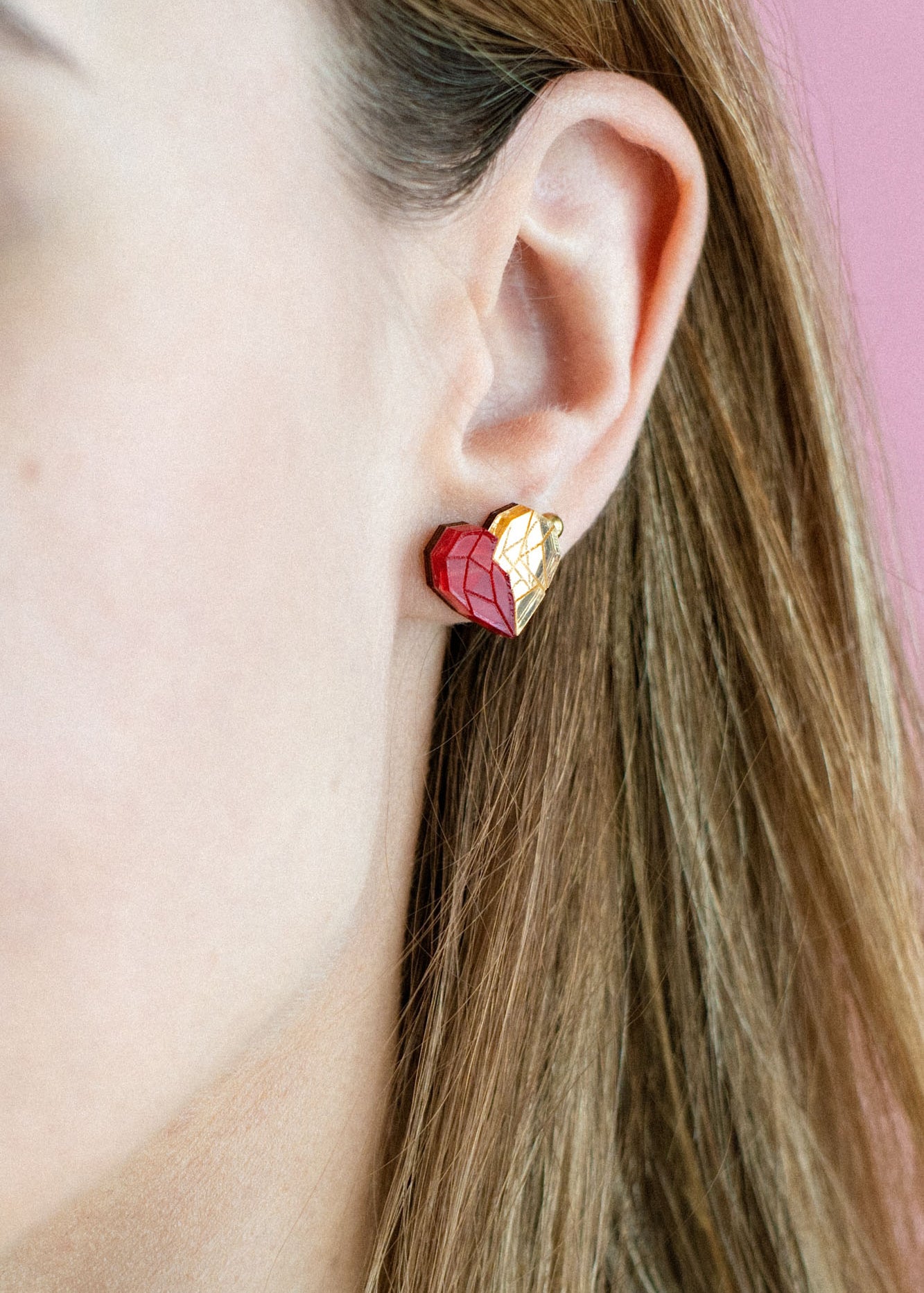 A close-up of a woman with light brown hair in a cream top, showing off romantic jewelry—a red and gold heart-shaped handcrafted earring. The pink background highlights the elegant design, with only the right side of her face visible.