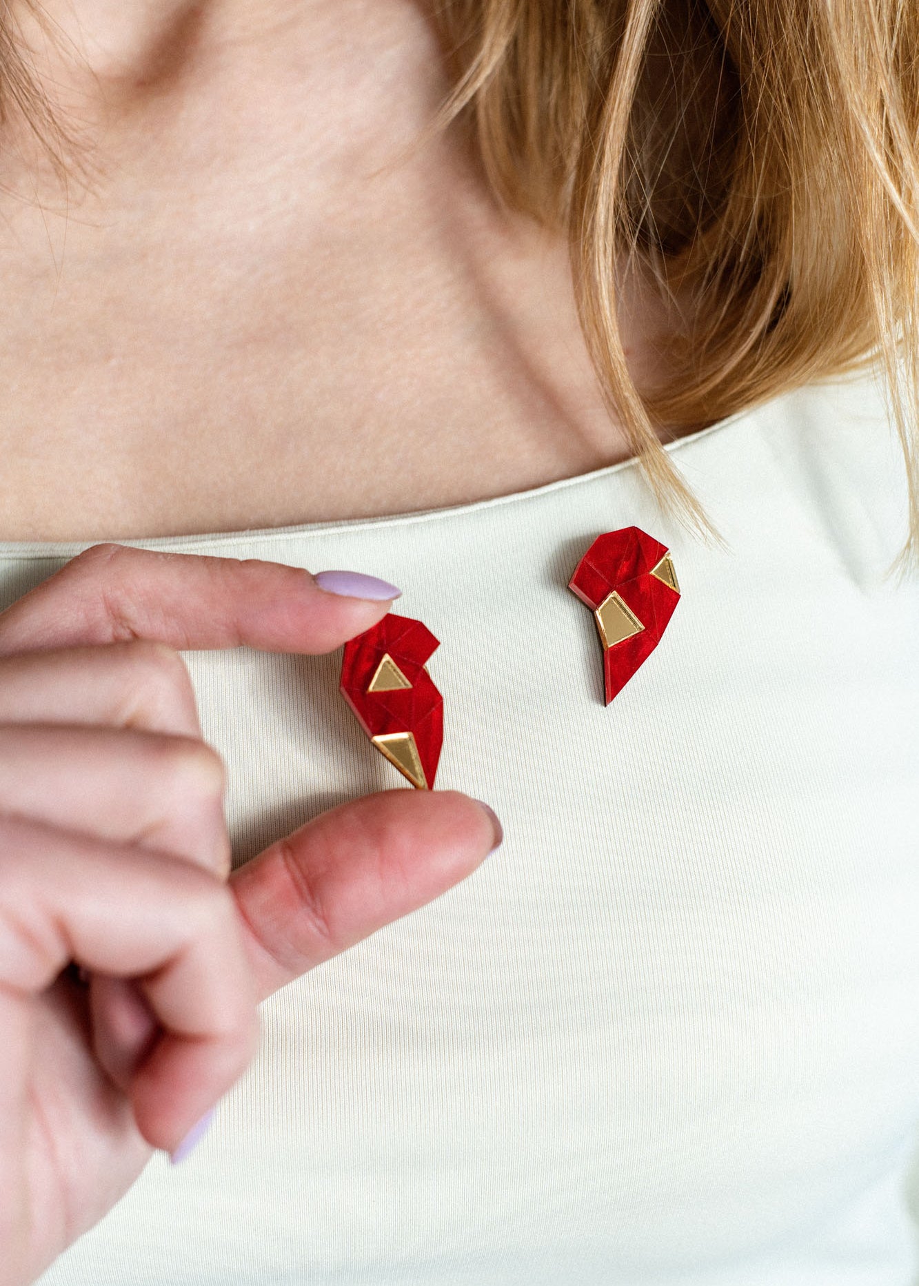A person with long hair wears a cream top, holding a red and gold geometric One Love Halves Pin shaped like half a broken heart—the other handcrafted half is pinned to their shirt near the collarbone. Perfect as a romantic gift.