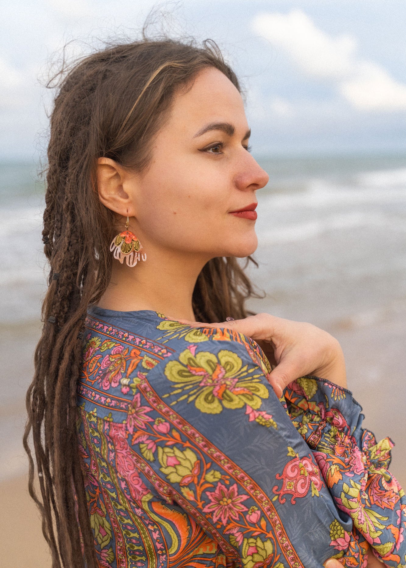 A woman with long, brown dreadlocks stands by the ocean, wearing a colorful, patterned shawl and Organic Floral Hoops accented with acrylic flowers, looking thoughtfully into the distance beneath a partly cloudy sky.