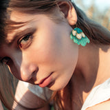 A young woman with long brown hair gazes intently at the camera, wearing a white top and handcrafted earrings—Organic Floral Hoops with turquoise and gold leaf accents. Natural outdoor lighting highlights her face.