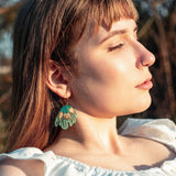 A young woman with light brown hair and bangs, wearing a white top and handcrafted earrings, stands outdoors in sunlight with her eyes closed, appearing peaceful.