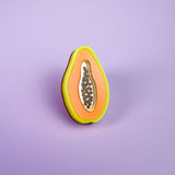 A handcrafted cookie decorated to look like a sliced papaya, with neon orange, yellow, and green icing plus black seeds, stands upright on a purple surface with a purple background—just like a Papaya Pin come to life.