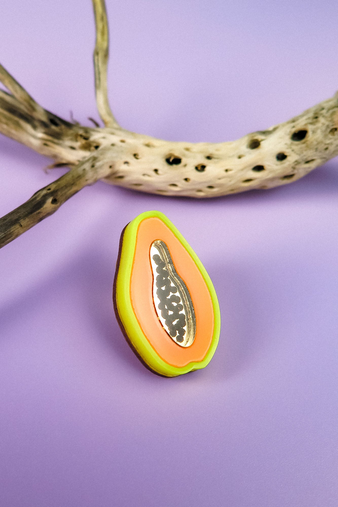 A handcrafted, colorful papaya-shaped cookie with icing details sits on a purple surface next to a dried, spotted branch, reminiscent of a neon Papaya Pin.