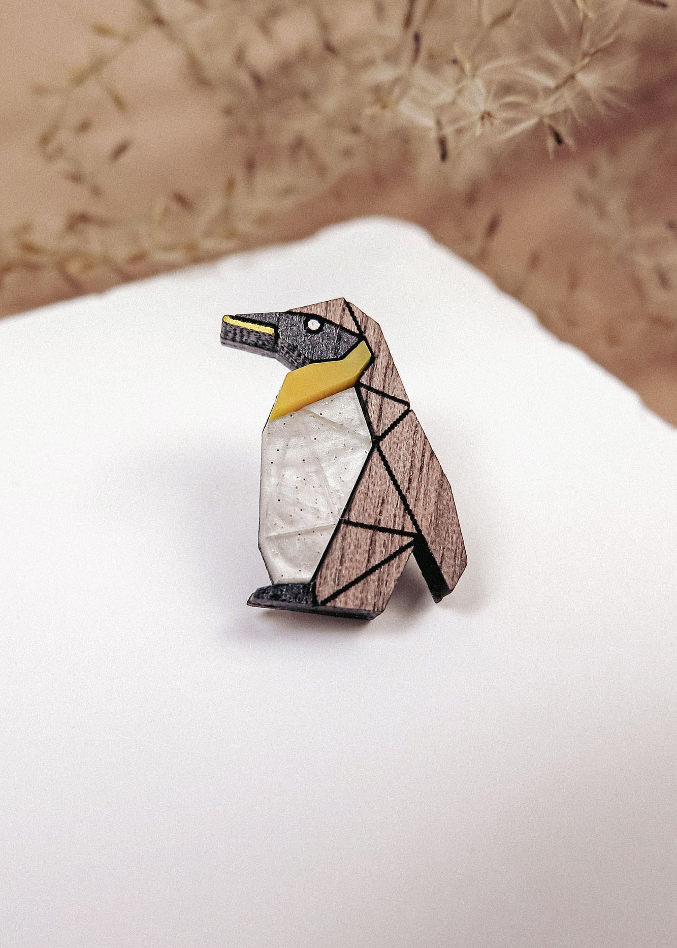 A geometric Penguin Pin accessory, crafted from wood in black, white, and yellow sections, rests on a white surface. Wispy beige plants are blurred in the background, making this brooch a charming addition to any outfit.