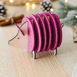 A decorative hedgehog figurine, crafted from wood and vibrant purple felt discs, sits on a light wooden table—an ideal housewarming gift—surrounded by festive lights, pinecones, and cinnamon sticks blurred in the background.