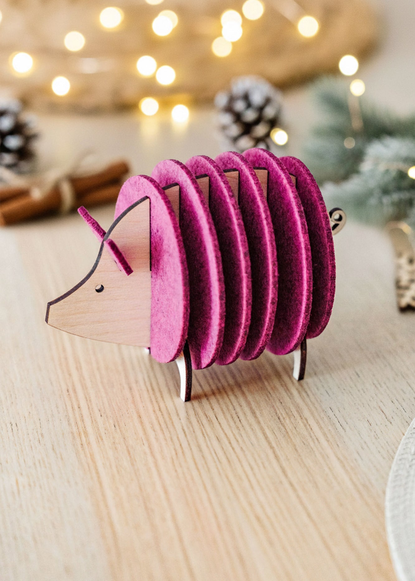 A decorative hedgehog figurine, crafted from wood and vibrant purple felt discs, sits on a light wooden table—an ideal housewarming gift—surrounded by festive lights, pinecones, and cinnamon sticks blurred in the background.