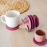 A white cup and a pink cup sit on purple felt coasters next to a wooden hedgehog-shaped coaster holder, with more felt coasters inside it, on a light wooden table—a lovely housewarming gift. A woven mat is in the background.
