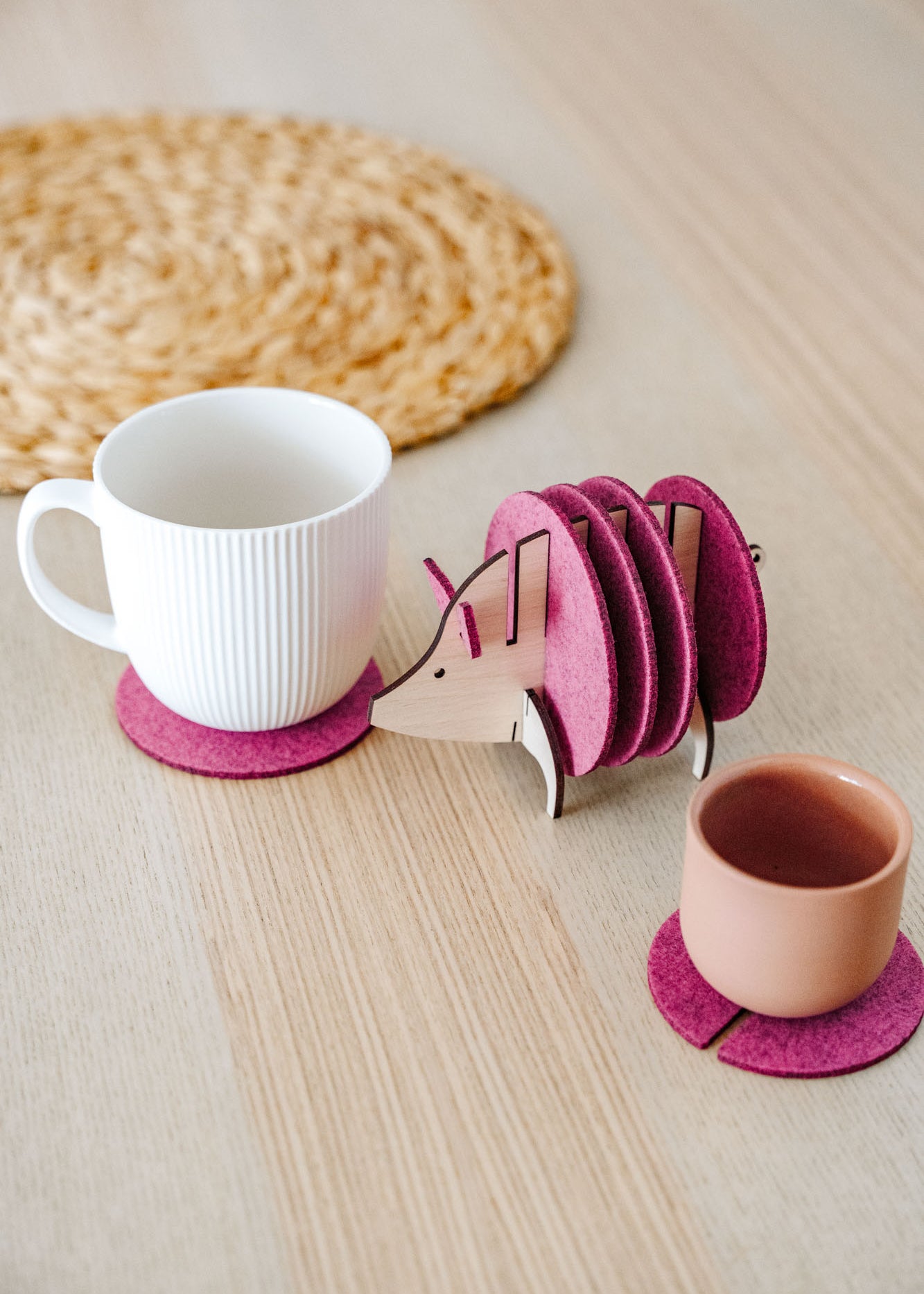 A white cup and a pink cup sit on purple felt coasters next to a wooden hedgehog-shaped coaster holder, with more felt coasters inside it, on a light wooden table—a lovely housewarming gift. A woven mat is in the background.
