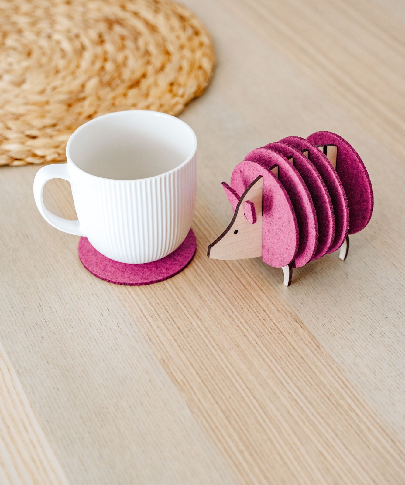 A white ribbed mug on a pink felt coaster sits next to a pink, hedgehog-shaped coaster holder on a light wooden table. A woven placemat is partially visible—perfect as a charming housewarming gift.