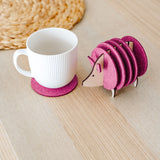 A white ribbed mug on a pink felt coaster sits next to a pink, hedgehog-shaped coaster holder on a light wooden table. A woven placemat is partially visible—perfect as a charming housewarming gift.