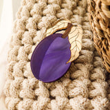 A purple enamel eggplant brooch with gold-outlined leaves rests on a chunky, cream-colored knit blanket beside a woven basket—crafted by a family business.