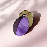 A decorative brooch shaped like a plum with two green leaves, crafted from acrylic and recycled wood, rests on a pale pink surface with soft shadows.