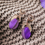 A pair of elegant earrings with purple circular charms and gold leaf accents rest on a textured beige knit fabric, showcasing the beauty of handcrafted earrings.
