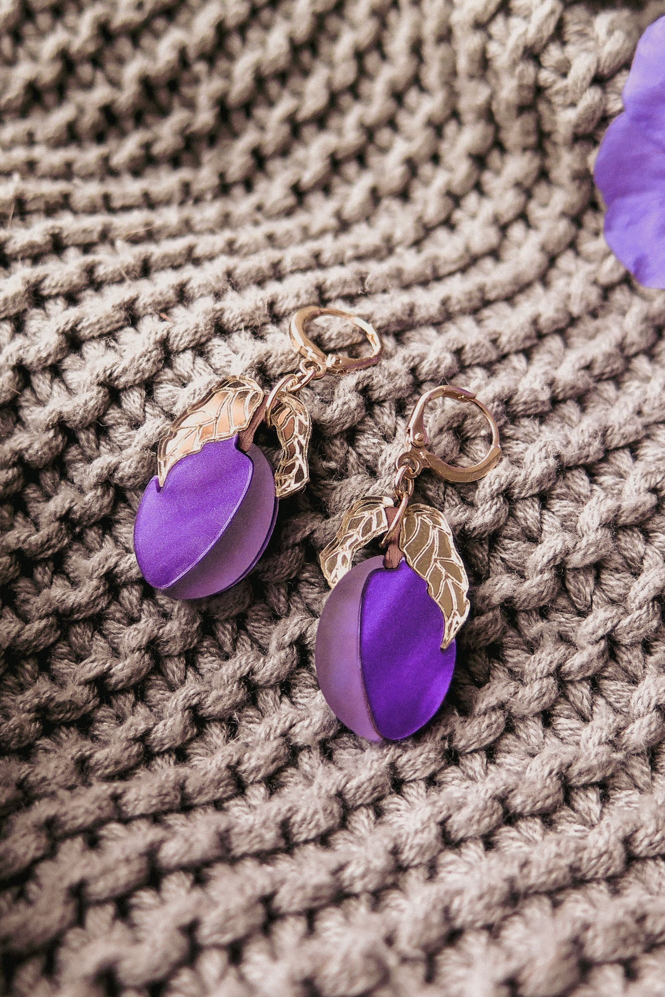 A pair of elegant earrings with purple circular charms and gold leaf accents rest on a textured beige knit fabric, showcasing the beauty of handcrafted earrings.