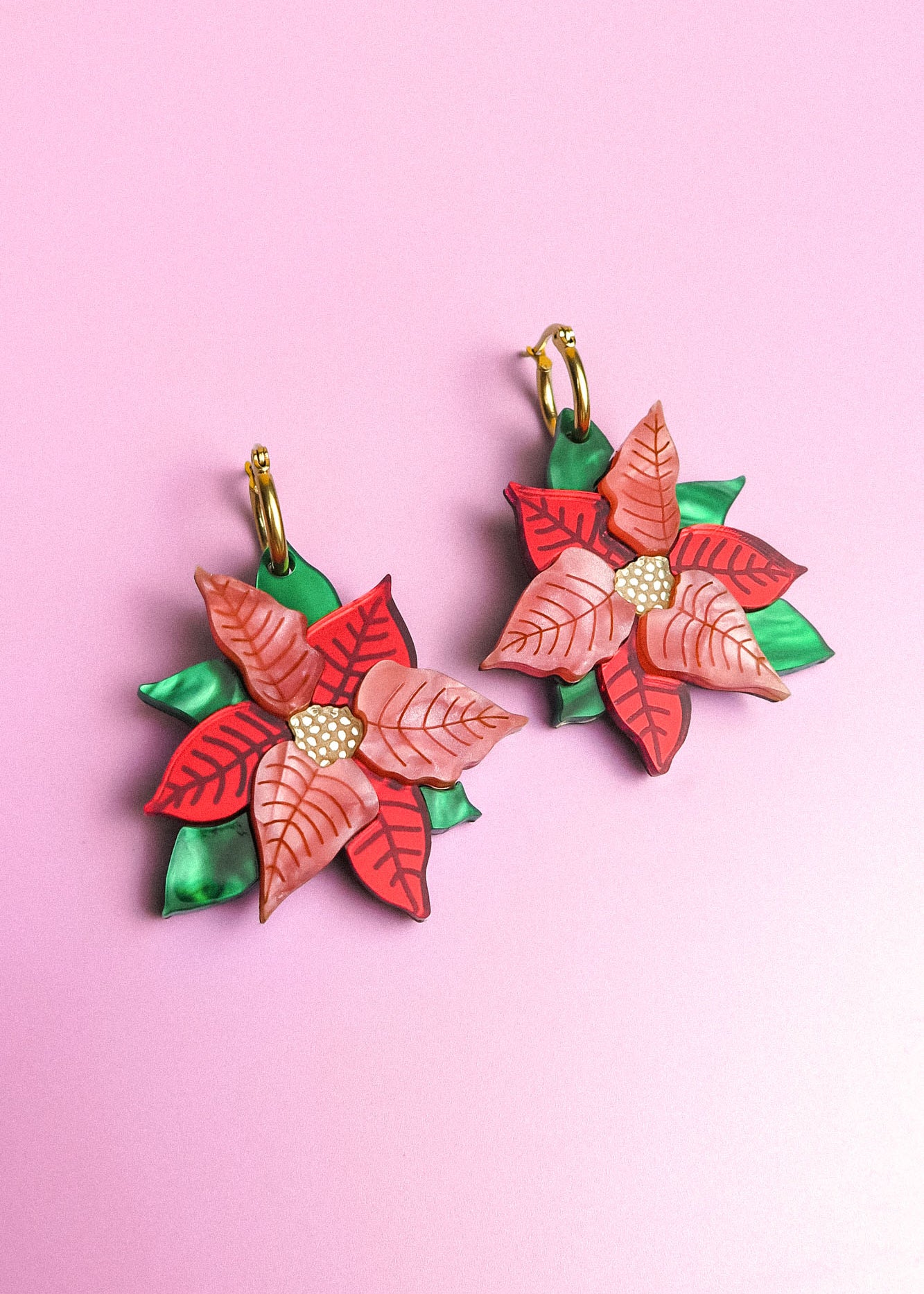 A pair of Poinsettia Statement Hoops shaped like poinsettia flowers with red and pink petals, yellow centers, and green leaves, displayed on a light purple background—perfect festive earrings for your holiday jewelry collection.