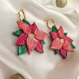 A pair of handmade Poinsettia Statement Hoops shaped like pink and red flowers with gold centers and green leaves, displayed on white satin fabric with two glittery gold decorative balls—perfect festive earrings for holiday jewelry lovers.