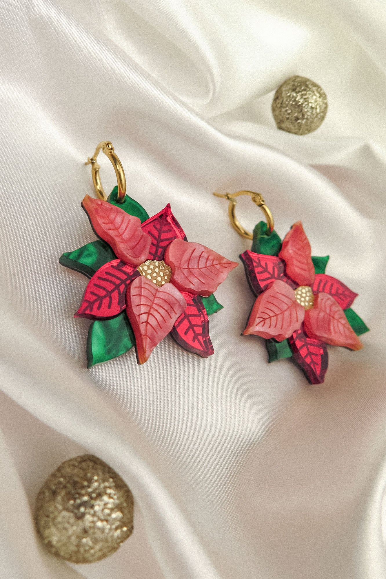 A pair of handmade Poinsettia Statement Hoops shaped like pink and red flowers with gold centers and green leaves, displayed on white satin fabric with two glittery gold decorative balls—perfect festive earrings for holiday jewelry lovers.