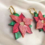 Close-up of festive earrings called Poinsettia Statement Hoops, featuring red and pink petals, green leaves, and gold hoop hooks, placed on white satin with a small gold ornament—perfect holiday jewelry for the season.