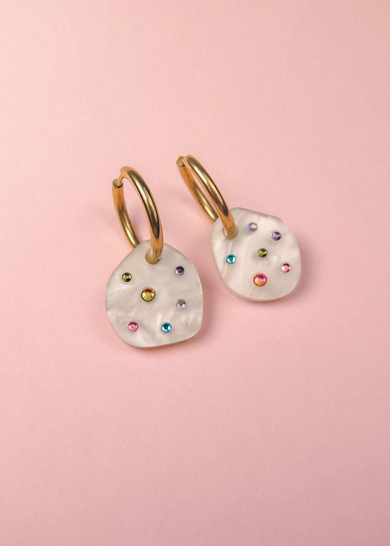 A pair of lightweight hoop earrings with irregular white, shell-like pendants, each adorned with small, colorful gemstone studs, displayed on a light pink background.