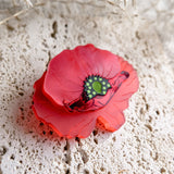 A detailed, red brooch shaped like a poppy with a green and black center rests on a textured, light-colored stone surface, its acrylic glass petals catching the light amid delicate dried grass in the background.