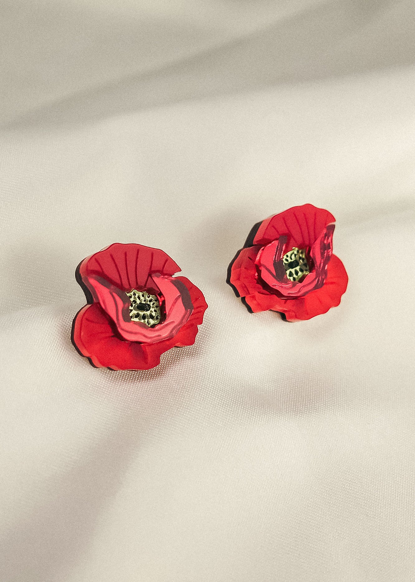 A pair of red, poppy design acrylic earrings with black and green centers, displayed on a soft, light-colored fabric background. Lightweight and eye-catching for any occasion.