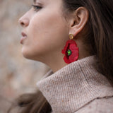 A woman wearing a beige turtleneck sweater is shown in profile. She has long brown hair and wears handcrafted Red Poppy Statement Earrings, a bold accessory with green and black details. The background is soft and out of focus.