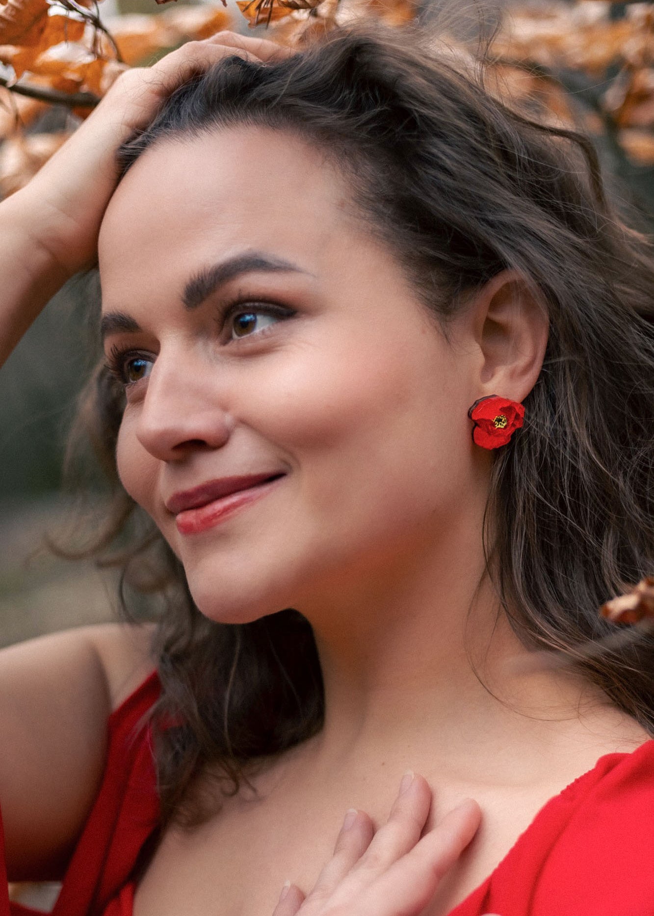 A woman with wavy brown hair, wearing a red dress and lightweight acrylic earrings with a poppy design, stands outdoors among autumn leaves, smiling softly with one hand on her head and the other on her chest.