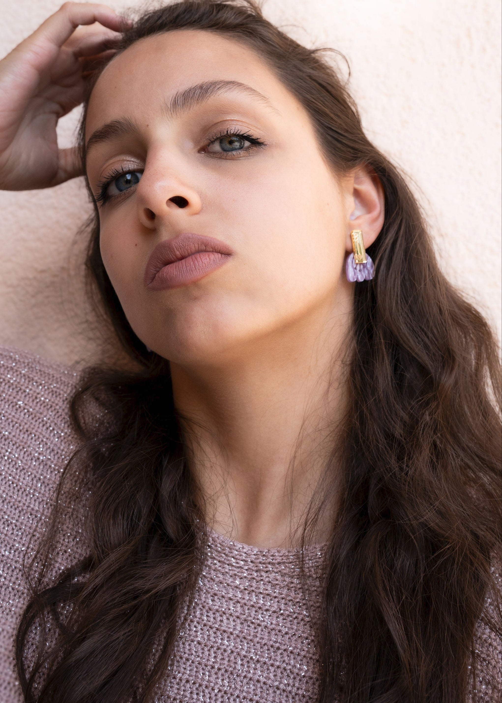 A woman with long brown hair wearing a sparkly pink sweater and handcrafted earrings looks confidently at the camera against a light background. Her hand rests on the side of her head, showing off her elegant accessory.