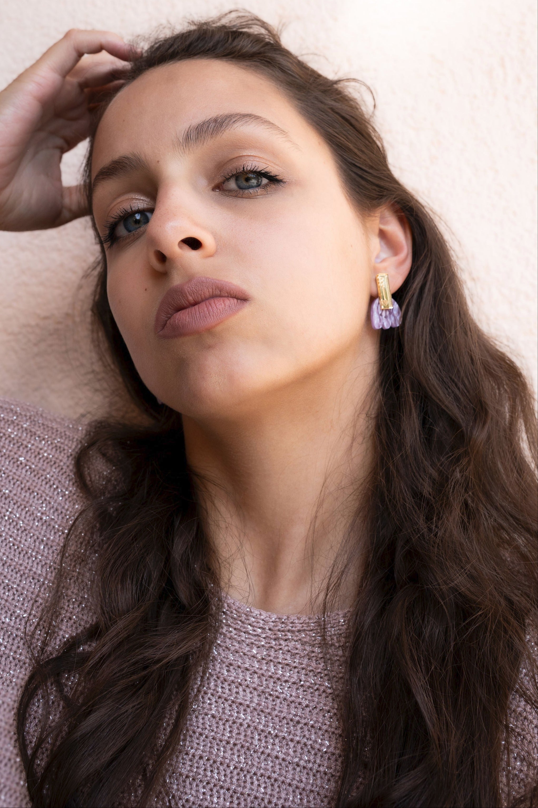 A woman with long brown hair wearing a sparkly pink sweater and handcrafted earrings looks confidently at the camera against a light background. Her hand rests on the side of her head, showing off her elegant accessory.