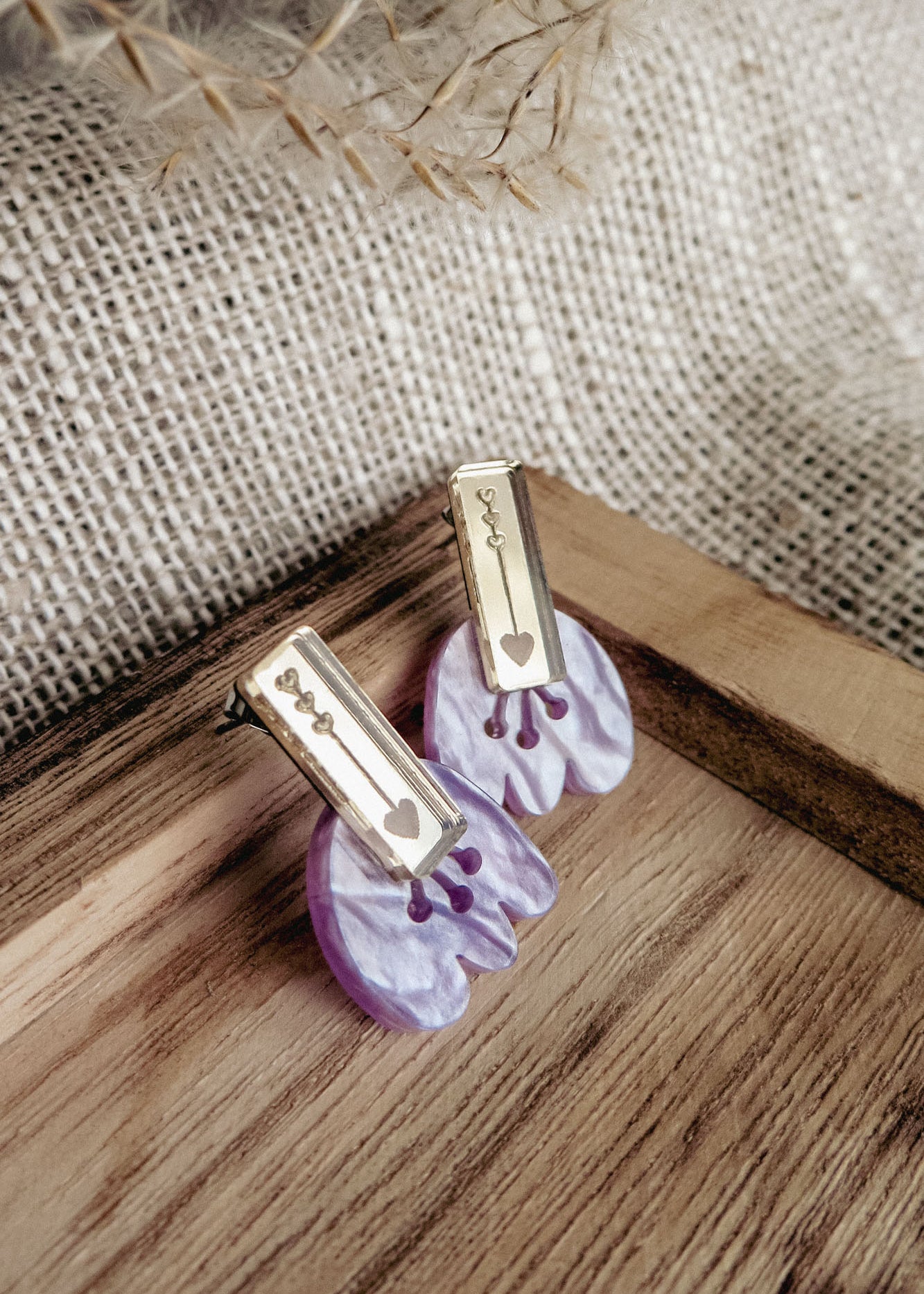 Romantic Tulip Earrings featuring gold rectangular tops engraved with a heart and abstract shapes, attached to marbled purple acrylic flower-shaped bases, offer allergy-free comfort. Displayed on a wooden tray with a burlap background.