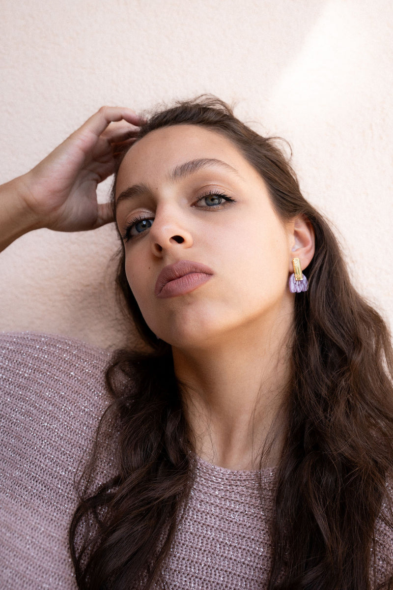 A woman with long brown hair and light eyes gazes confidently at the camera, wearing mauve lipstick, a textured mauve sweater, and Romantic Tulip Earrings in marbled purple acrylic for allergy-free comfort, her hand resting on her head.