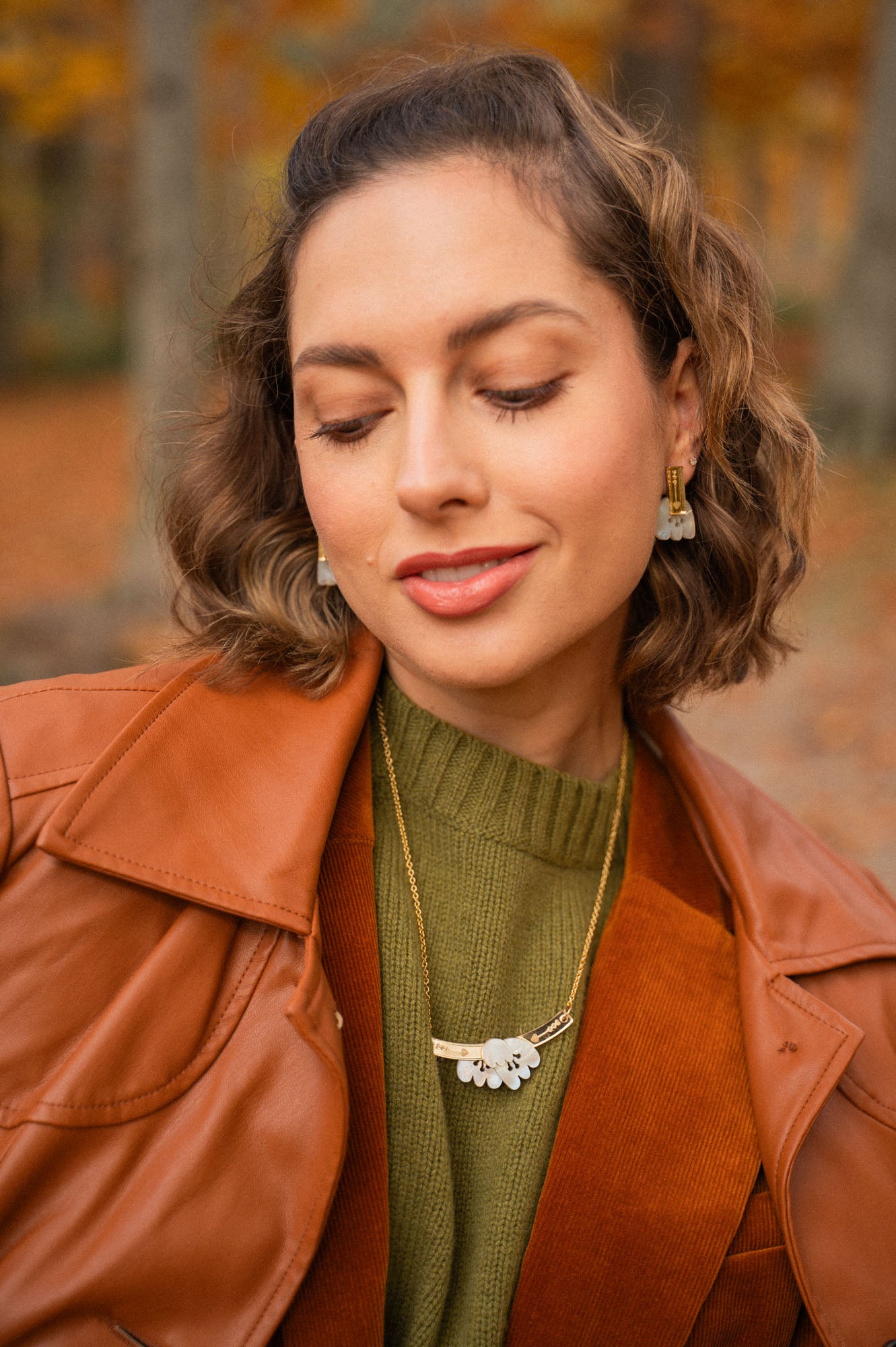 A woman with wavy brown hair, wearing a rust-colored jacket, olive green sweater, gold necklace with a floral pendant, and handcrafted Romantic Tulip Earrings, smiles gently with her eyes closed against an autumnal outdoor background.