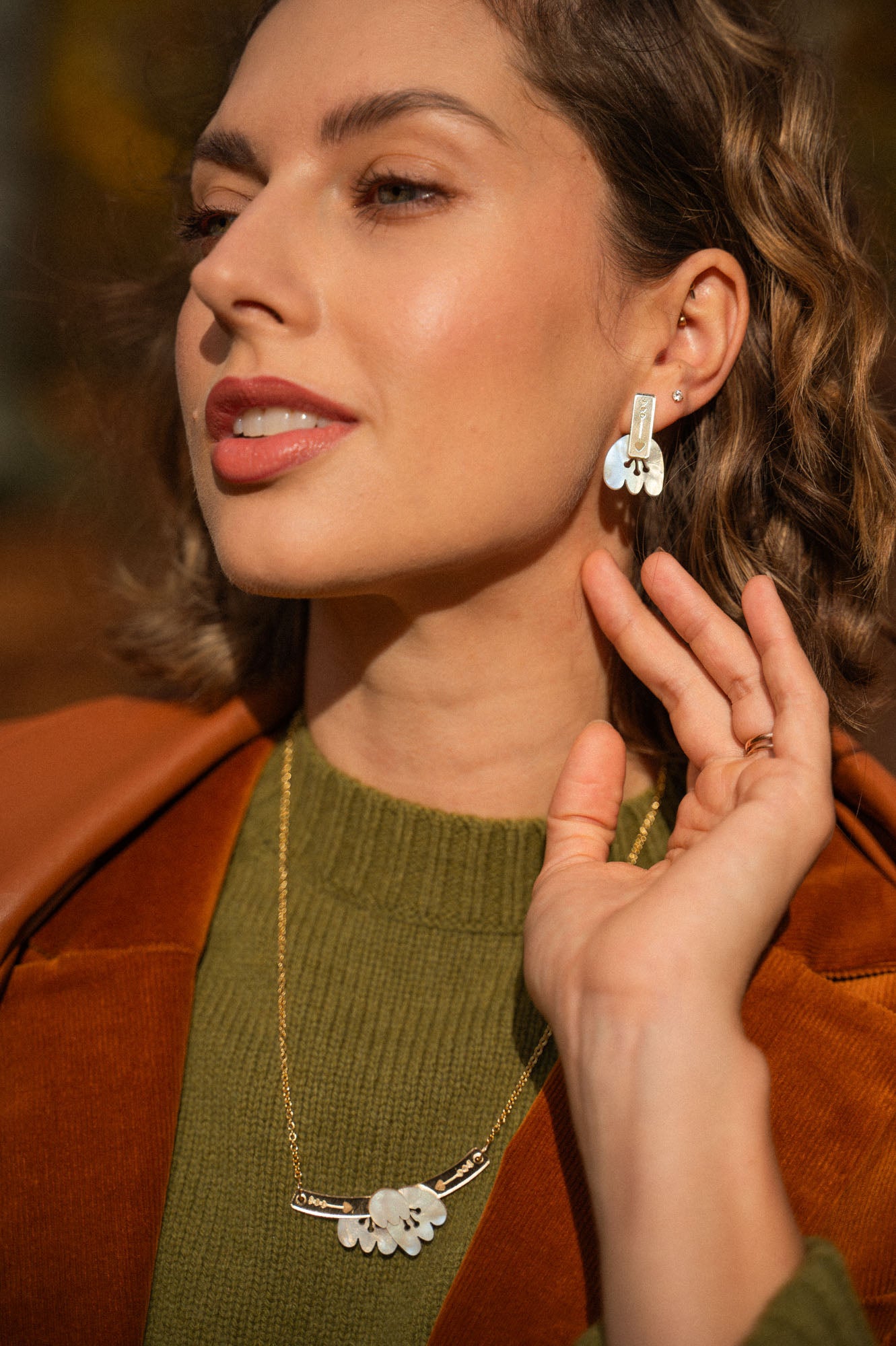 A woman with wavy, shoulder-length hair wears a green sweater, brown jacket, and gold handcrafted earrings—Romantic Tulip Earrings—with a matching necklace, gently touching her ear and smiling softly in warm sunlight.