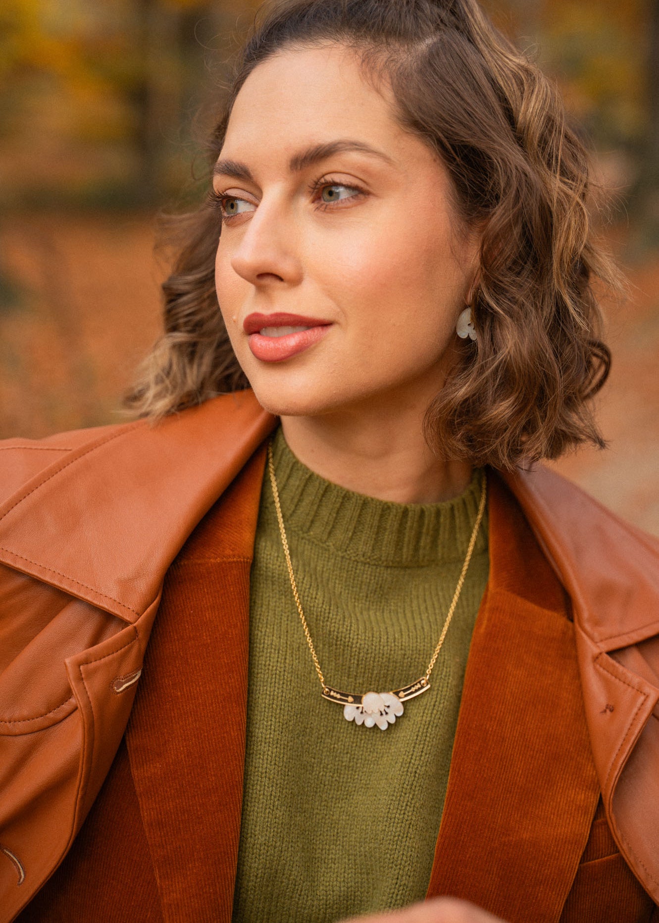 A woman with short, wavy brown hair wears a brown leather jacket over a green sweater and an elegant Tulip Necklace with a white floral pendant, standing outdoors with an autumn background.