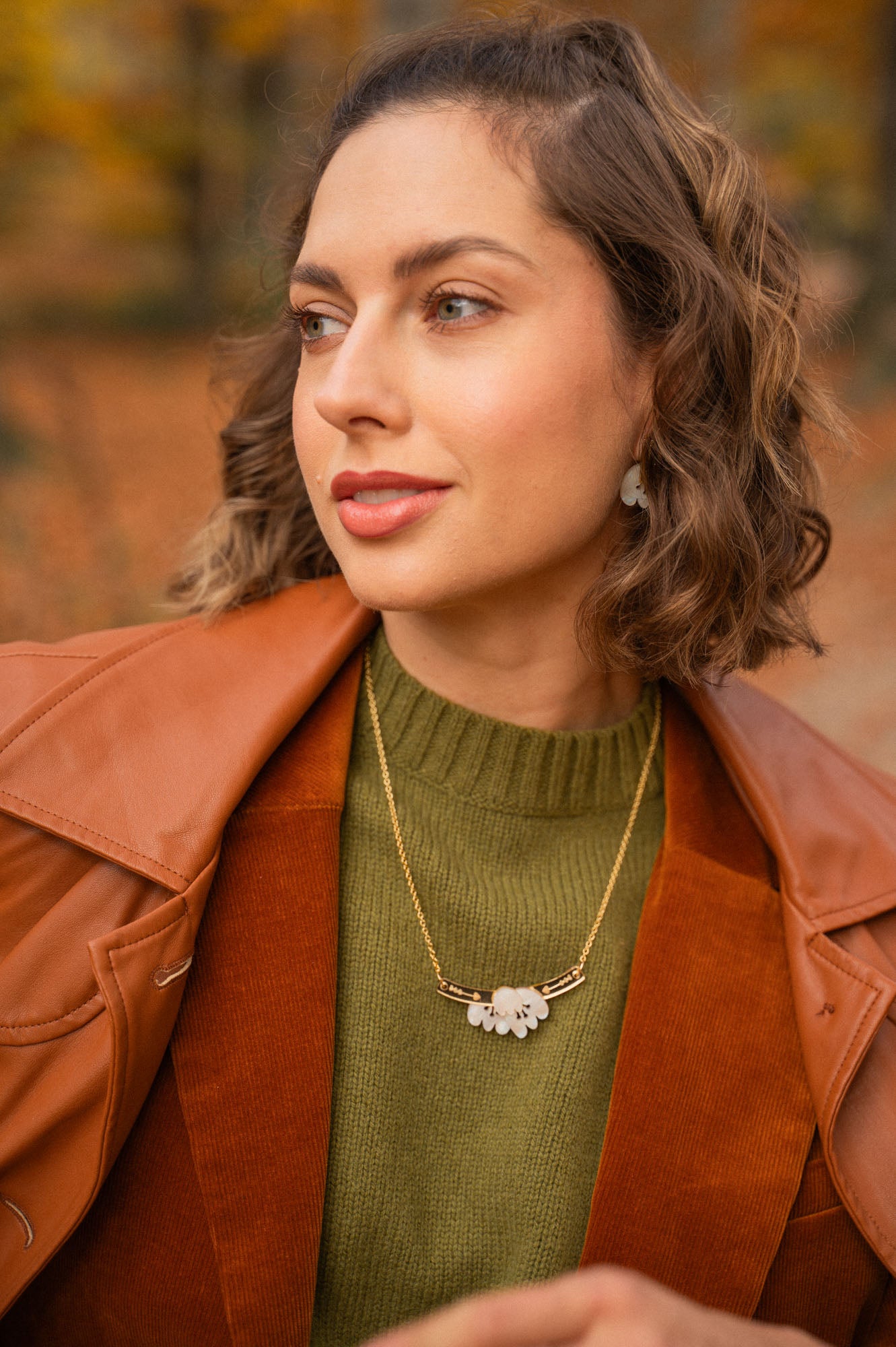 A woman with short, wavy brown hair wears a brown leather jacket over a green sweater and an elegant Tulip Necklace with a white floral pendant, standing outdoors with an autumn background.