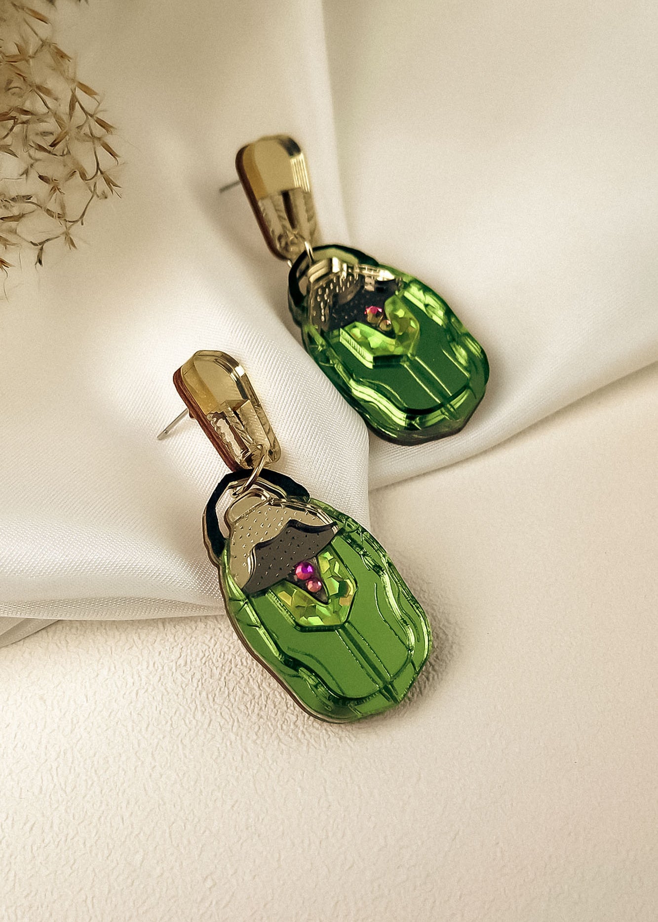 A pair of handcrafted gold and green Rose Chafer Earrings with an abstract face design, featuring purple and black accents, displayed on a cream-colored fabric background with dried flowers in the corner.