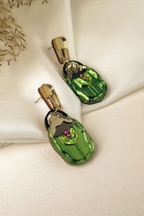 A pair of handcrafted gold and green Rose Chafer Earrings with an abstract face design, featuring purple and black accents, displayed on a cream-colored fabric background with dried flowers in the corner.