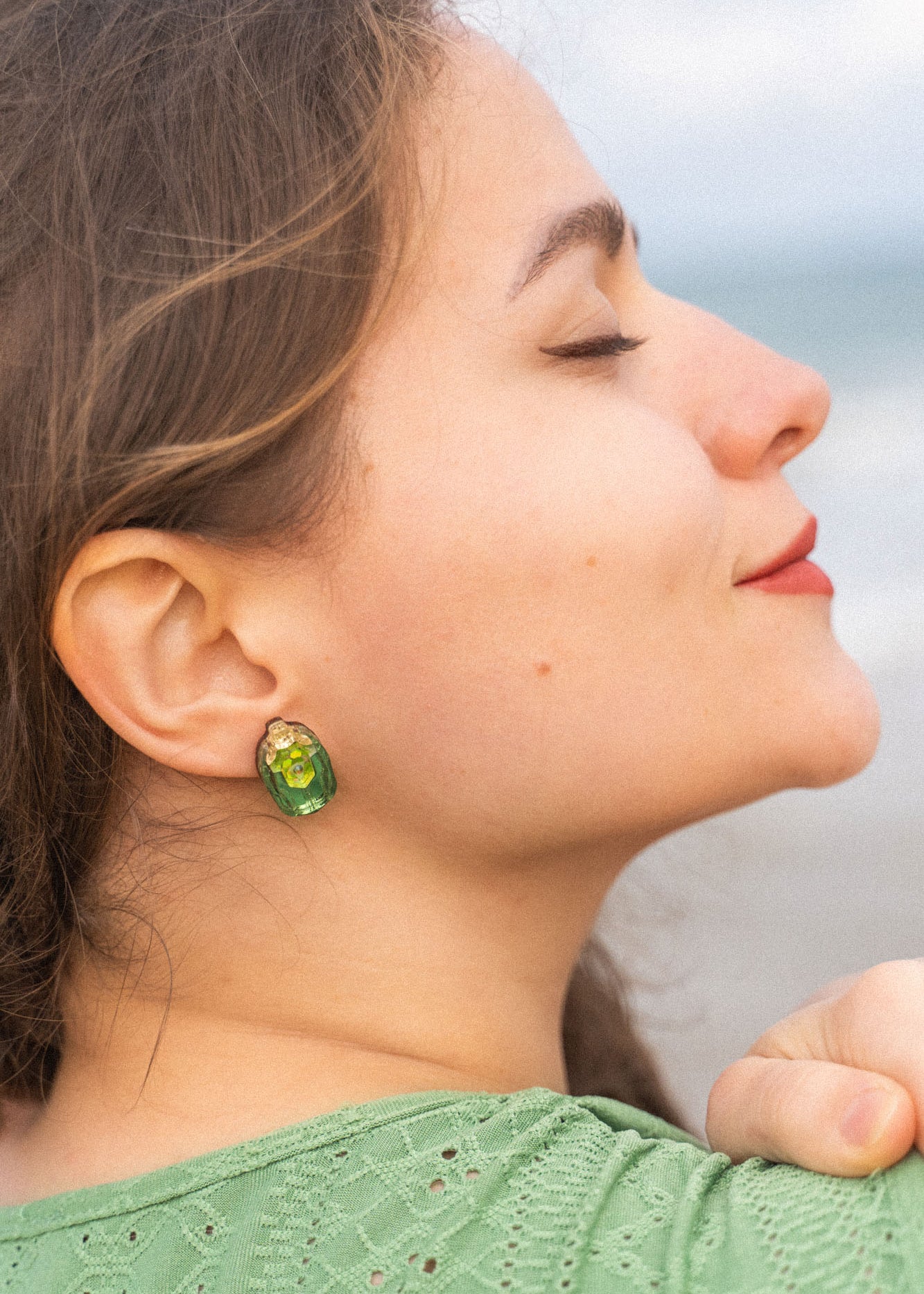 A woman with closed eyes and a slight smile faces sideways, wearing a green top and nature-inspired earrings, with the beach and ocean blurred in the background.