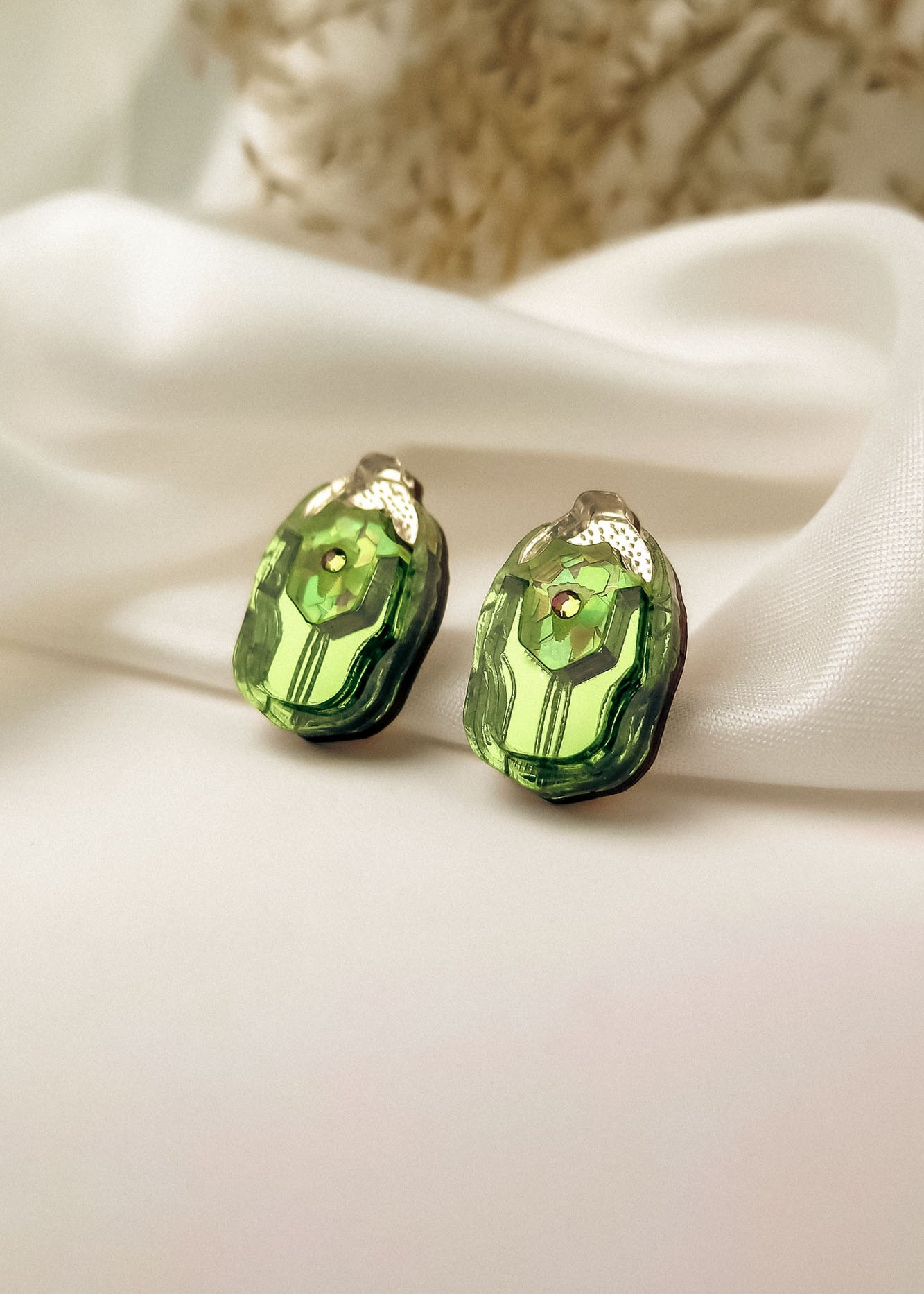 A pair of green, faceted gemstone Rose Chafer Studs with metallic tops displayed on white satin fabric, these nature-inspired earrings shine against a soft, blurred background.