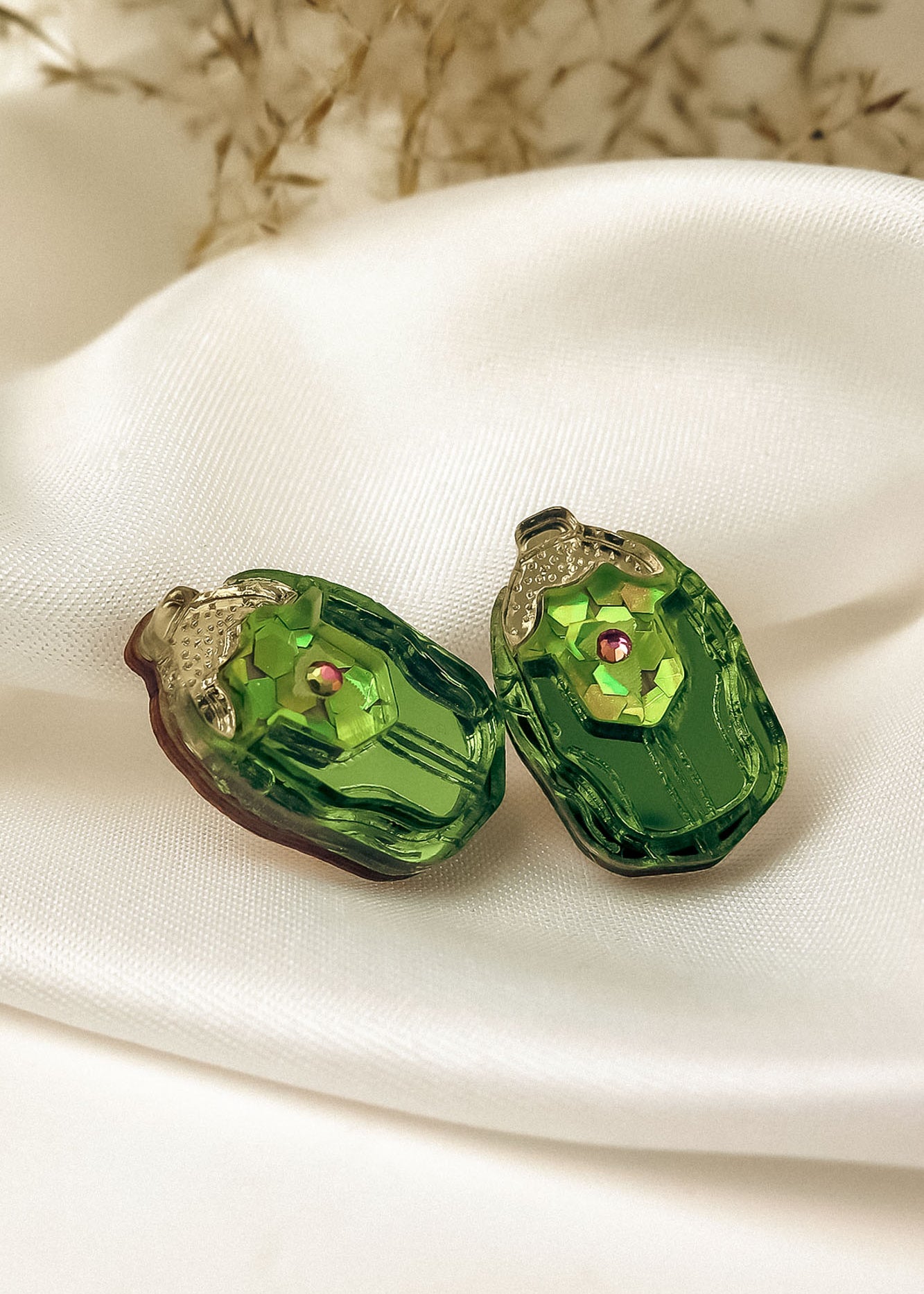 Two green, oval-shaped glass craft beads with a faceted surface and a small pink center gem, perfect for nature-inspired earrings, resting on a white silky fabric backdrop with blurred dried flowers in the background.
